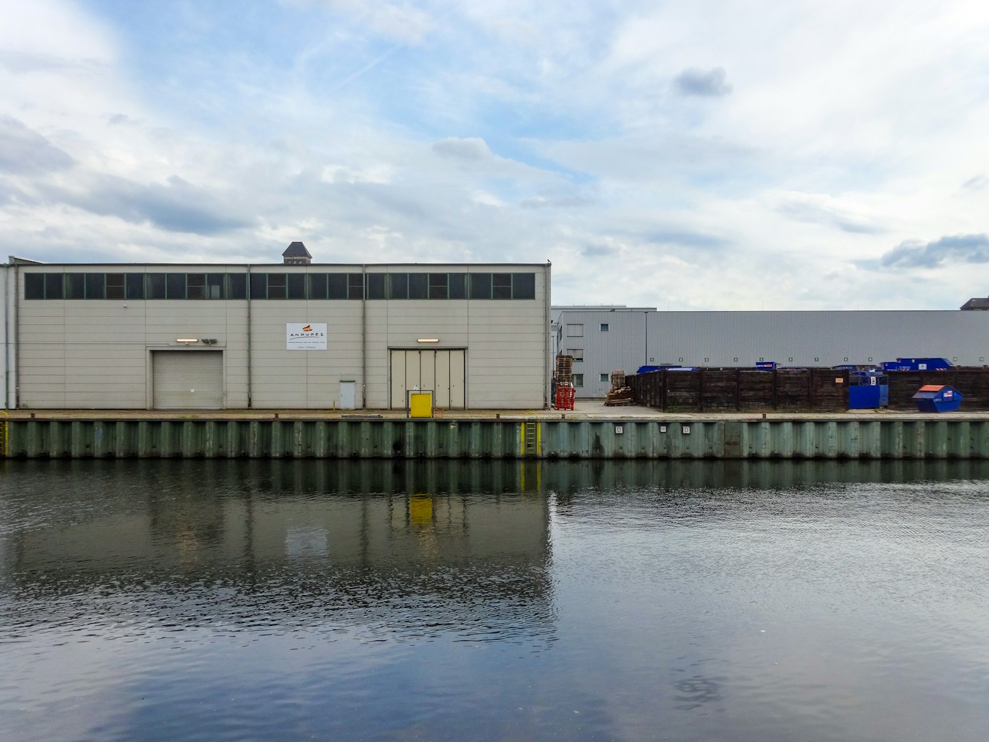 Industriegebäude reihen sich an ein ruhiges Ufer unter grauem Himmel.